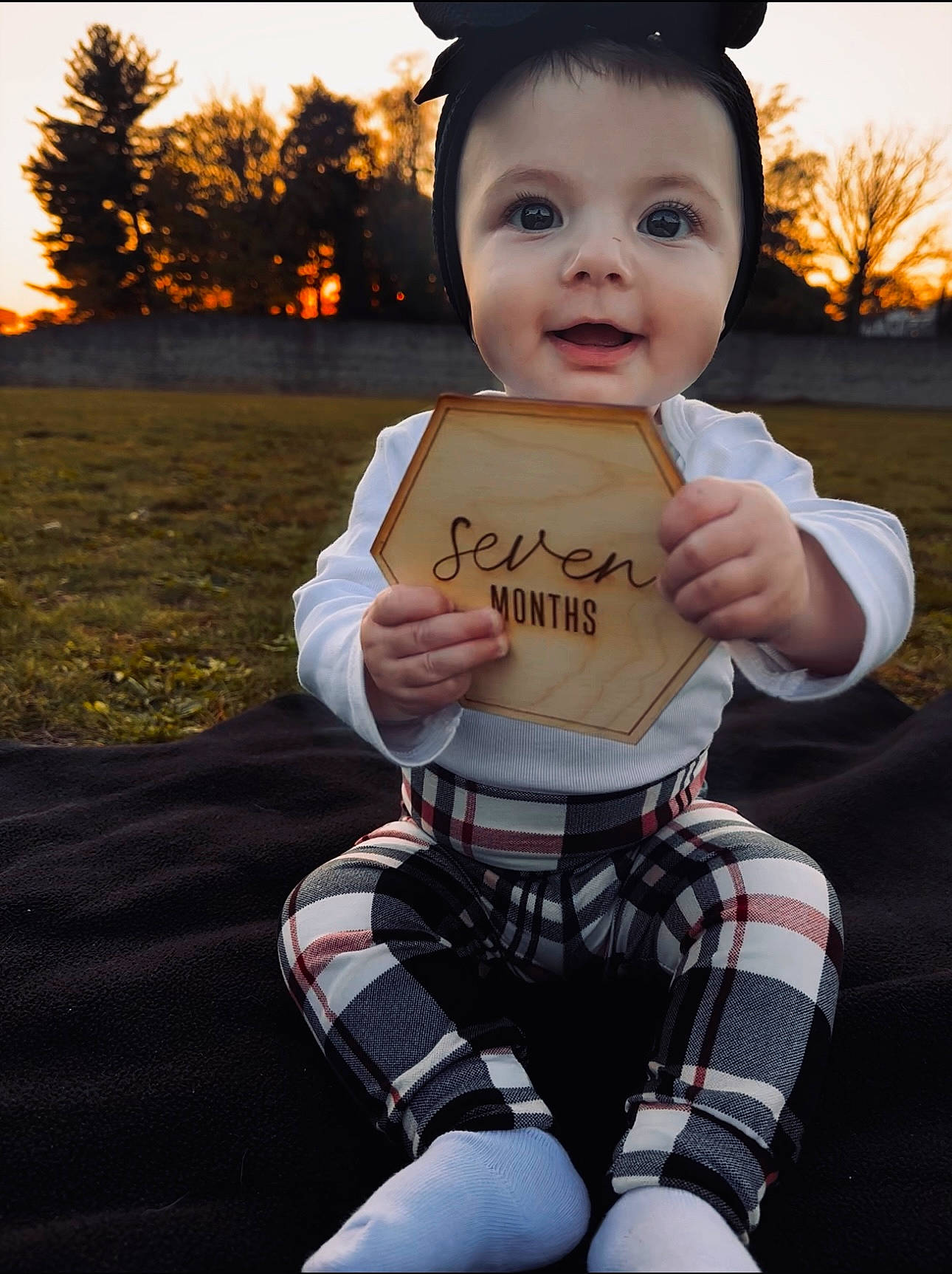 Myli is registered to the contest to win money with this photo: baby, child, coffee_cup, gesture, grass, happy, hat, human_leg, joy, knee, pattern, person, personal_protective_equipment, plaid, recreation, sitting, sky, smile, tartan, toddler