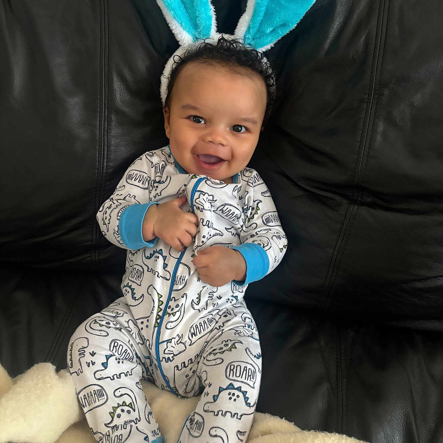 Kasaan is registered to the contest to win money with this photo: baby, black_leather_couch, blanket, blue, bunny_ears, child, cozy, curly_hair, cute, dinosaur_print, happy, headband, indoors, infant, onesie, playful, portrait, sitting, smiling, white