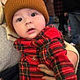 baby, child, plaid_clothing, knit_hat, red_clothing, brown_hat, person, hand, face, curious_expression, indoor, people, blurred_background, holding, warm_clothing, cute, portrait, young_child, infant, closeup