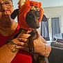 woman, dog, glasses, orange_hat, costume, indoor, living_room, tattoo, holding, pet, brown_dog, short_hair, furniture, curtains, casual_clothing, expression, face, hand, portrait, cute