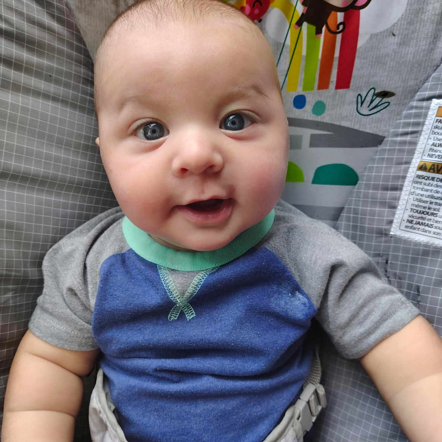 Oryon is registered to the contest to win money with this photo: baby, baby_seat, blue_eyes, blue_shirt, child, chubby_cheeks, close_up, colorful_background, cute, gray_clothing, happy, human, indoors, infant, monkey_illustration, person, portrait, rainbow, safety_strap, smiling