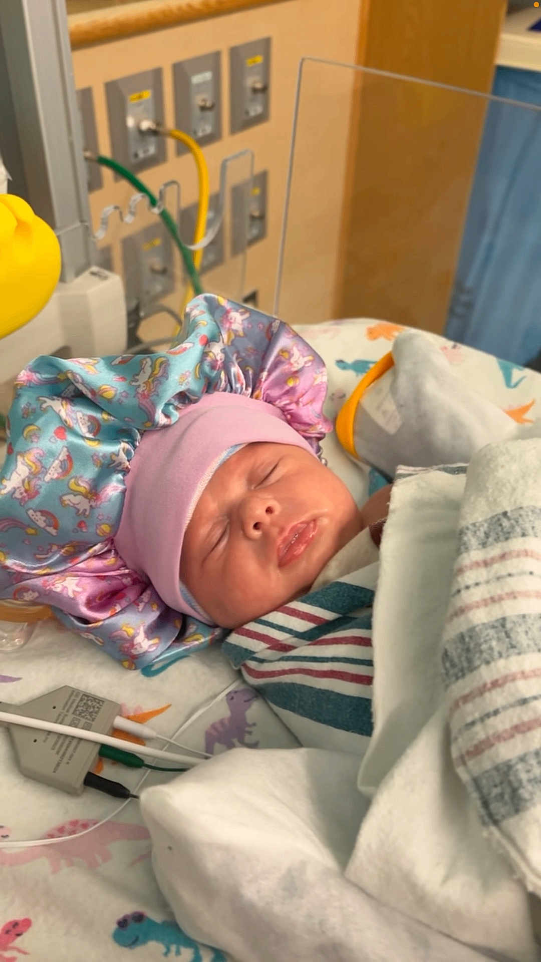 Aleiah is registered to the contest to win money with this photo: newborn, baby, sleeping, hospital, blanket, bonnet, unicorn, rainbow, medical_equipment, wires, dinosaur_print, bed, face, clothing, infant, cute, resting, child, wrapped, indoors