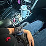 air_freshener, arm, black_cat, car_interior, car_seat, cat, clothing, collar, console, cup, dashboard, hand, jeans, kitten, mug, person, seatbelt, sleeping, steering_wheel, travel_mug