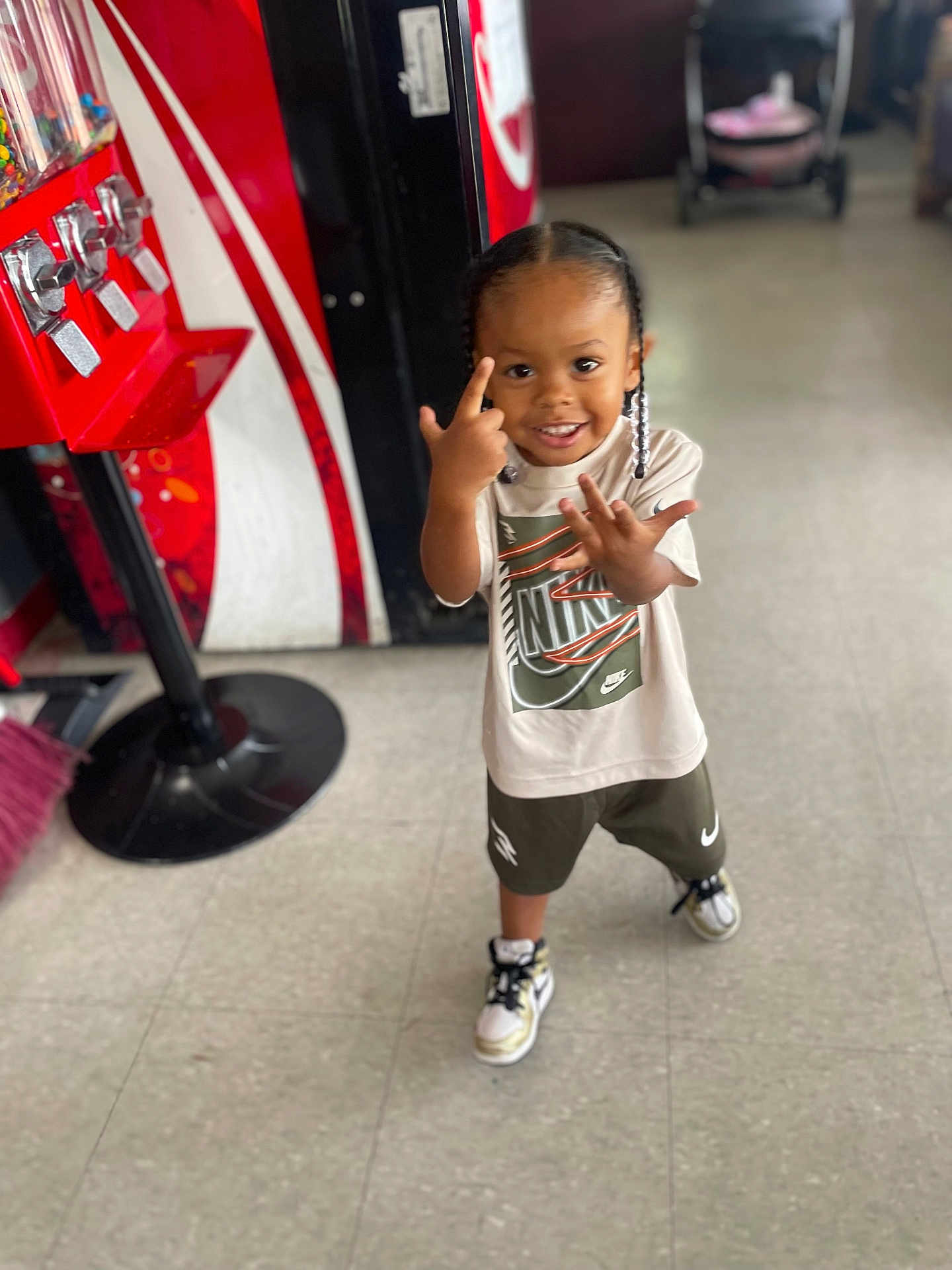 Keyontae is registered to the contest to win money with this photo: child, smiling, braids, indoor, nike, sneakers, tiled_floor, candy_dispenser, stroller, clothing, person, happy, young_child, standing, playful, casual_wear, footwear, retail, floor, portrait