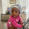 adult_hand, baby, bonnet, child, curious, dark_skin, earrings, headband, indoor, infant, kitchen, long_sleeves, looking_at_camera, mickey_mouse, pink_clothing, portrait, sitting, table, toy, window_blinds