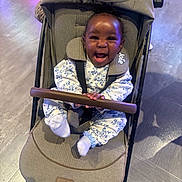 Leilani is registered to the contest to win money with this photo: baby, child, stroller, smiling, happy, portrait, indoor, floral_outfit, white_clothing, safety_harness, seated, infant, face, hands, feet, floor, lighting, colorful, joyful, baby_carriage