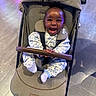 baby, child, stroller, smiling, happy, portrait, indoor, floral_outfit, white_clothing, safety_harness, seated, infant, face, hands, feet, floor, lighting, colorful, joyful, baby_carriage