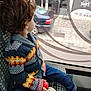 child, toddler, curly_hair, knitted_sweater, colorful_clothing, blue_jeans, white_shoes, red_ball, window, bus_seat, public_transport, side_view, urban_scene, car_outside, patterned_seat, indoor, daylight, casual, sitting, looking_out