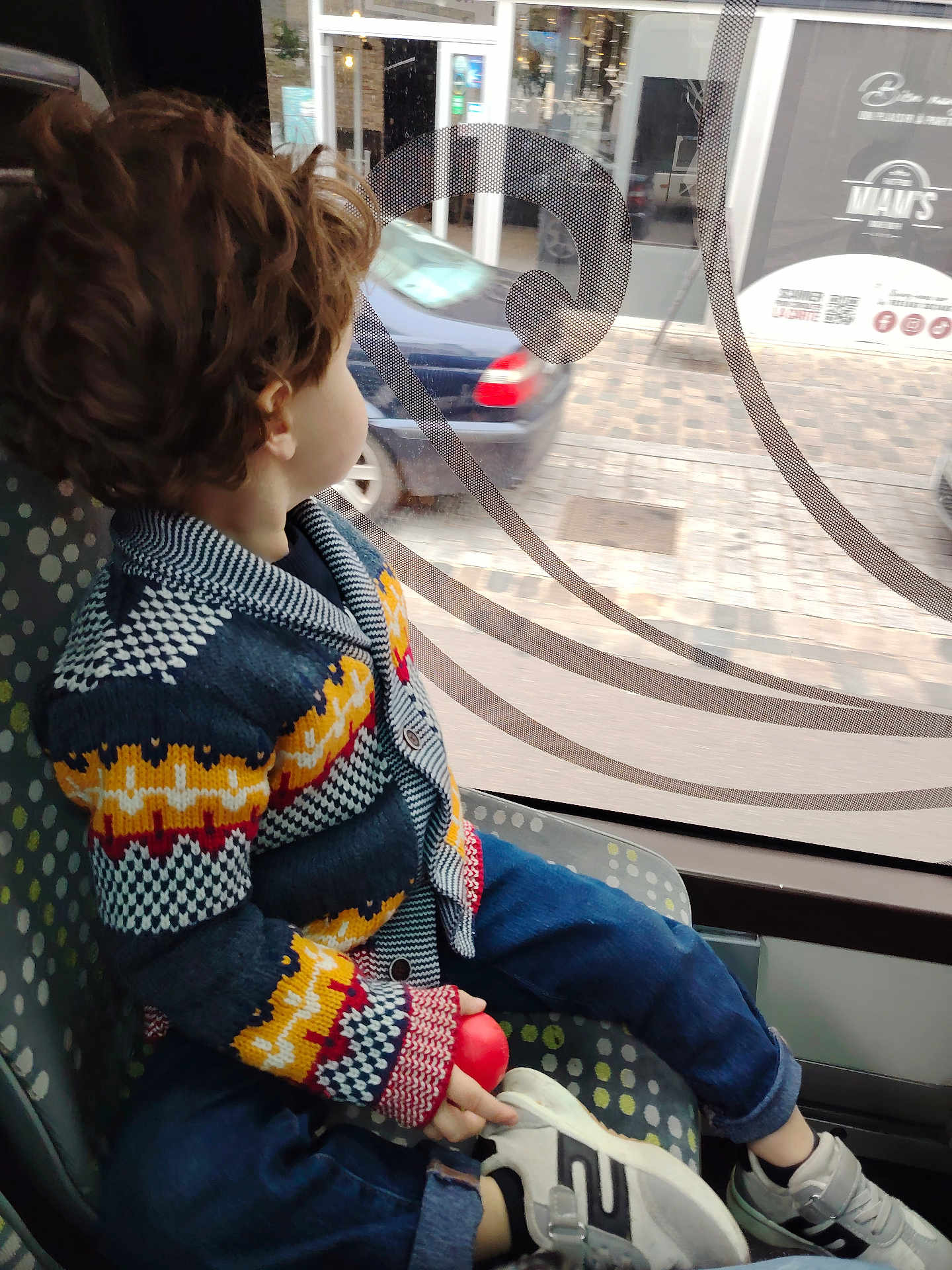 O H a rejoint le concours — aidez-le/la à gagner de superbes lots ! child, toddler, curly_hair, knitted_sweater, colorful_clothing, blue_jeans, white_shoes, red_ball, window, bus_seat, public_transport, side_view, urban_scene, car_outside, patterned_seat, indoor, daylight, casual, sitting, looking_out