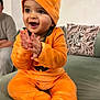 Célia a rejoint le concours — aidez-le/la à gagner de superbes lots ! baby, child, clapping, clothing, couch, cute, green_couch, happy, hat, home, indoor, joyful, orange_costume, person, pillows, plush, pumpkin, seated, smiling, toddler