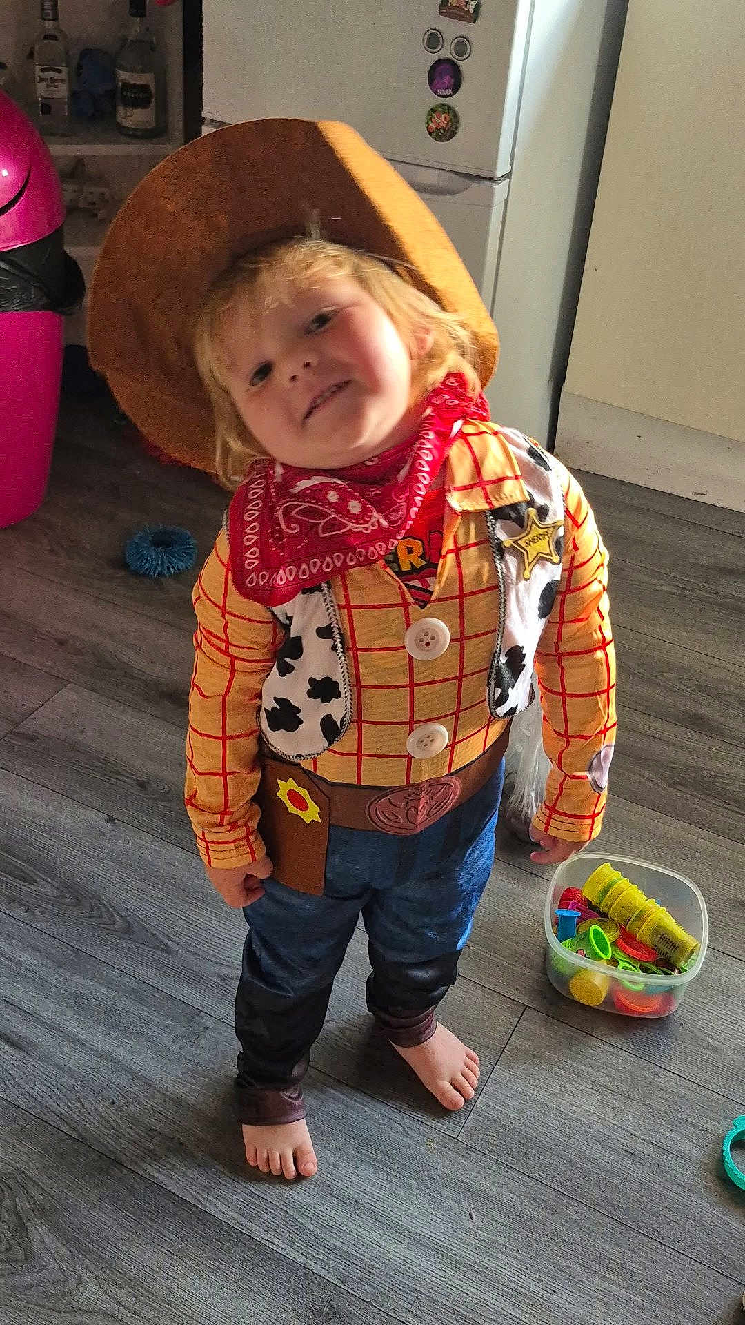 Bradley is registered to the contest to win money with this photo: child, toddler, costume, cowboy, hat, bandana, barefoot, indoor, floor, toy, plastic_container, fridge, wooden_floor, smiling, cute, clothing, play, home, person, sheriff_badge