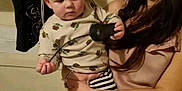Bradley is registered to the contest to win money with this photo: baby, bag, black_object, casual, child, cord, curious, curly_hair, floor, holding, home, indoor, patterned_shirt, person, portrait, shelf, striped_pants, stuffed_animals, wall, woman
