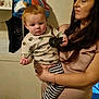 Bradley is registered to the contest to win money with this photo: baby, child, woman, indoor, holding, curious, striped_pants, patterned_shirt, stuffed_animals, bag, shelf, floor, wall, person, portrait, home, black_object, cord, casual, curly_hair