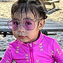 child, pink_jacket, watermelon_pattern, sunglasses, purple_lenses, braided_hair, beach, sand, picnic_table, wooden_bench, outdoor, daylight, portrait, casual_clothing, curious_expression, young_girl, summer, sunlight, close_up, fashion