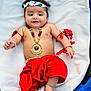 baby, child, smiling, traditional_clothing, red_skirt, jewelry, bracelet, headband, peacock_feather, lying_down, blanket, cute, happy, infant, anklet, cultural, portrait, indoors, decorative, celebration