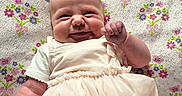 Evee joined the competition — help win amazing prizes! baby, infant, smile, dress, blanket, floral_pattern, cute, chubby_cheeks, lying_down, soft_lighting, indoors, happy, person, toddler, hand, face, skin, fabric, cozy, portrait