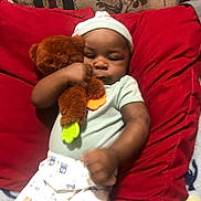 Keison joined the competition — help win amazing prizes! baby, baby_clothes, bed, blanket, brown_skin, child, cozy, cuddling, face, indoors, infant, onesie, pacifier, photograph, portrait, red_pillow, seated, sleepy, teddy_bear, toy