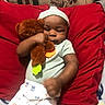 baby, baby_clothes, bed, blanket, brown_skin, child, cozy, cuddling, face, indoors, infant, onesie, pacifier, photograph, portrait, red_pillow, seated, sleepy, teddy_bear, toy