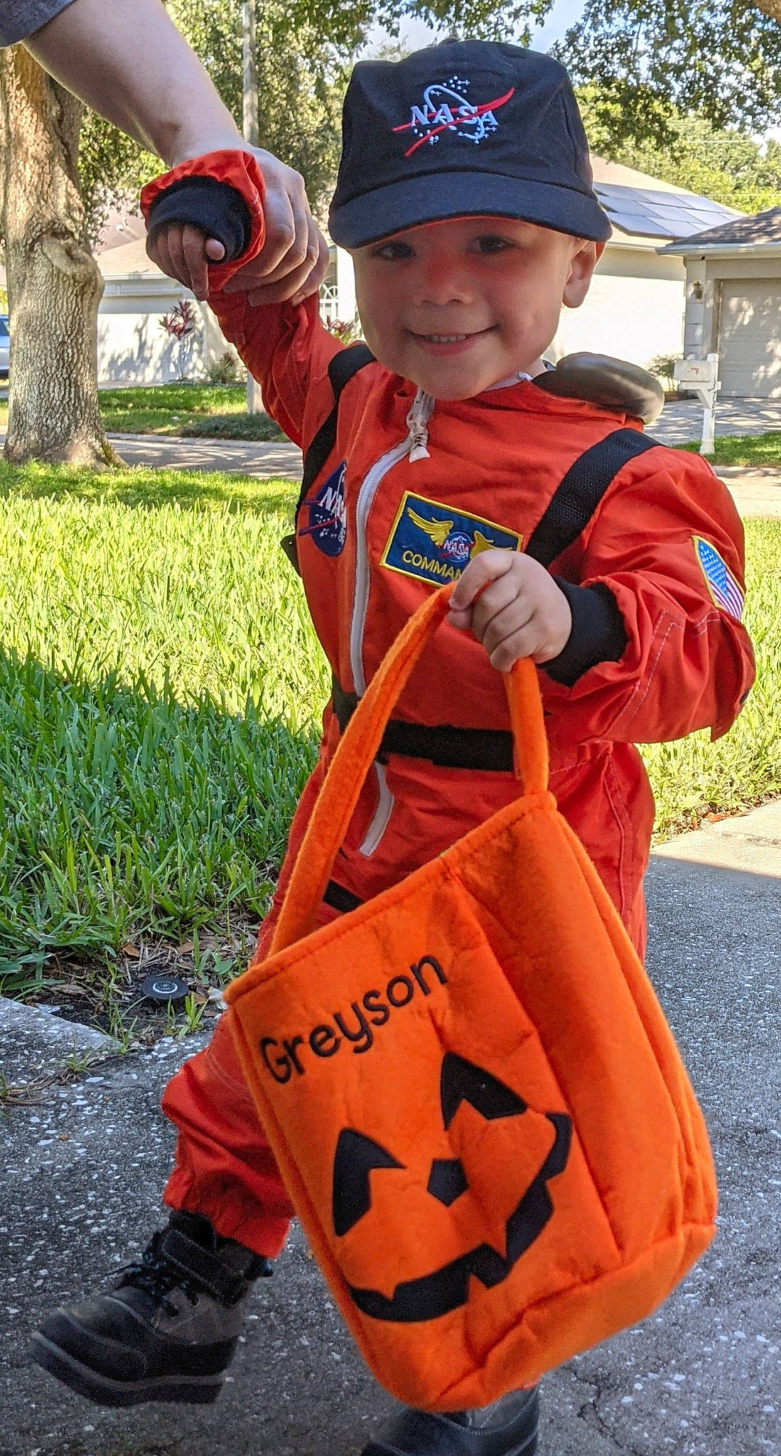 Greyson is registered to the contest to win money with this photo: bag, cap, fun, grass, green, headwear, high_visibility_clothing, joy, lifejacket, luggage_and_bags, orange, outerwear, person, personal_protective_equipment, plant, plastic_bag, red, sleeve, smile, toddler