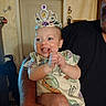 baby, child, crown, clapping, happy, smiling, person, indoor, adult_arm, shirt, face, home, light, door, furniture, joy, celebration, cute, closeup, portrait