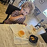 child, toddler, curly_hair, striped_shirt, eating, restaurant, table, chair, bread, dipping_sauce, drink, phone, video, indoor, tile_floor, window, bench, napkin, food, meal
