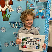 Samuel joined the competition — help win amazing prizes! child, smiling, curly_hair, sign, classroom, bulletin_board, pre_k, school, education, ocean_theme, decorations, happy, preschool, student, indoor, person, poster, colorful, young_child, front_view