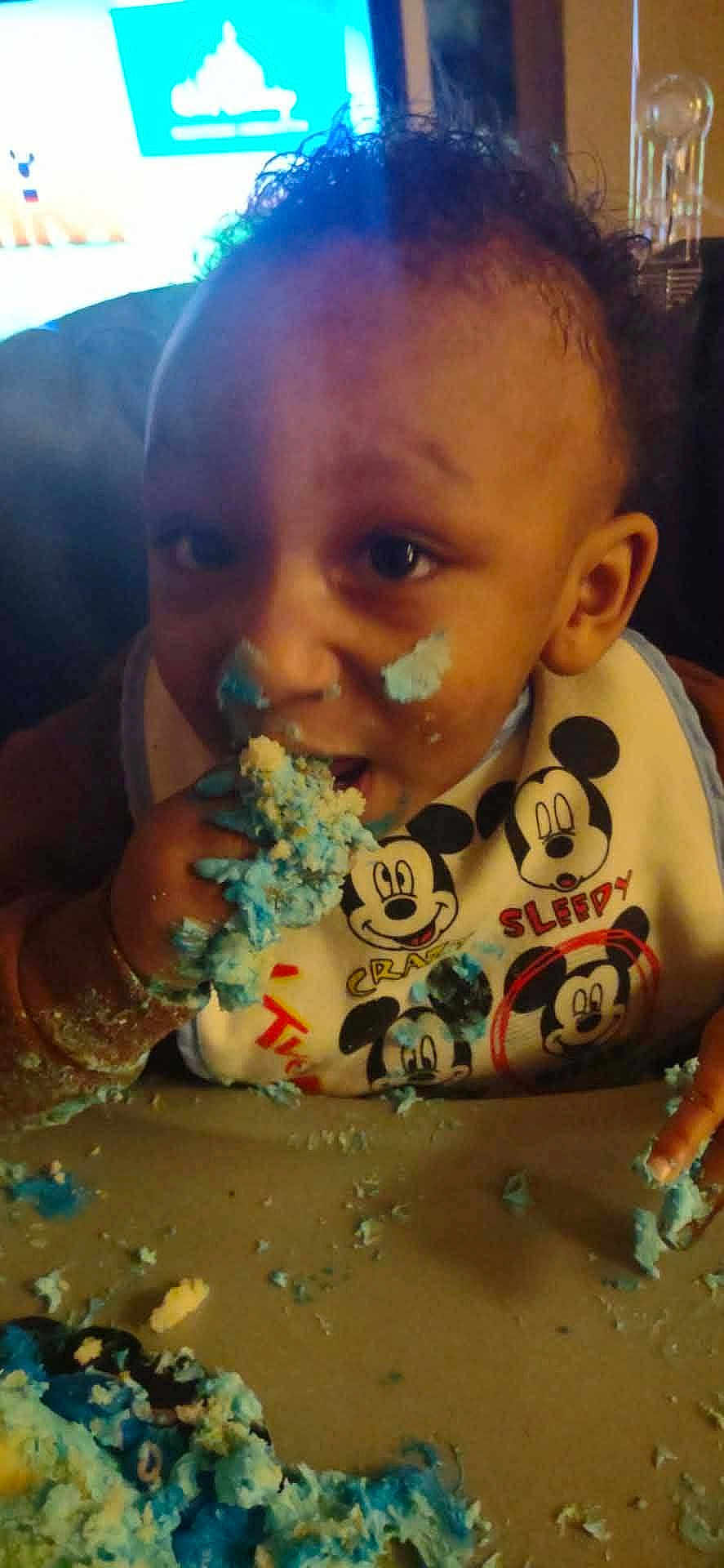Dominic joined the competition — help win amazing prizes! child, toddler, bib, mickey_mouse, cake, blue_frosting, messy, eating, hands, face, crumbs, highchair, tray, indoor, close_up, party, celebration, television, smile, expression
