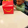 child, toddler, curly_hair, green_sweater, happy_meal_box, red_box, smile_logo, table, restaurant_booth, indoor, high_chair, hands, curious_expression, portrait, person, fast_food, seat, commercial_packaging, dining_table, meal_time