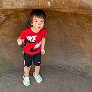 Grayson joined the competition — help win amazing prizes! child, boy, red_shirt, nike, shorts, rock, cave, outdoor, curious, expression, shadow, ground, sunlight, nature, footwear, clothing, person, standing, holding, small_object