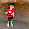 child, boy, red_shirt, nike, shorts, rock, cave, outdoor, curious, expression, shadow, ground, sunlight, nature, footwear, clothing, person, standing, holding, small_object