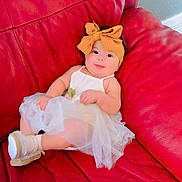 Dailany Abigail is registered to the contest to win money with this photo: baby, child, red_couch, orange_bow, headband, white_dress, footwear, leather, indoor, smiling, cute, infant, sitting, portrait, relaxed, furniture, skin, person, young_child, comfortable