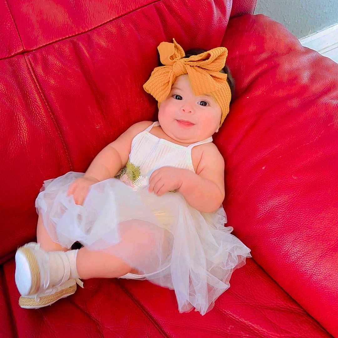 Dailany Abigail is registered to the contest to win money with this photo: baby, child, red_couch, orange_bow, headband, white_dress, footwear, leather, indoor, smiling, cute, infant, sitting, portrait, relaxed, furniture, skin, person, young_child, comfortable
