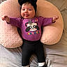 baby, infant, child, purple_shirt, black_bow, headband, pink_cushion, lying_down, tongue_out, black_leggings, white_socks, black_bow_shoes, bed, smiling, cute, indoors, soft_texture, playful, panda_graphic, peek_a_boo_text