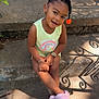 toddler, child, smiling, sitting, outdoor, steps, concrete, sunlight, shadow, hair_accessory, pink_slippers, bracelet, yellow_shirt, rainbow_print, happy, portrait, cute, daylight, casual_clothing, young_girl