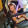 baby, stroller, balloon, purple_balloon, pink_hat, smiling, indoor, child, person, hand, foot, clothing, toy, wheel, carpet, bag, striped_bag, adult, black_stroller, happy