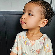 Sarai joined the competition — help win amazing prizes! child, toddler, portrait, curly_hair, profile, face, eyes, ear, looking_away, calm, cute, seated, shirt, seashell_pattern, starfish_pattern, ocean_theme, indoor, tile_background, dark_seat, close_up