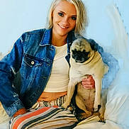 Missy is registered to the contest to win money with this photo: woman, dog, pug, denim_jacket, striped_pants, smiling, sitting, couch, indoor, pet, casual, blonde_hair, relaxed, portrait, human, animal, friendship, comfort, happy, cozy