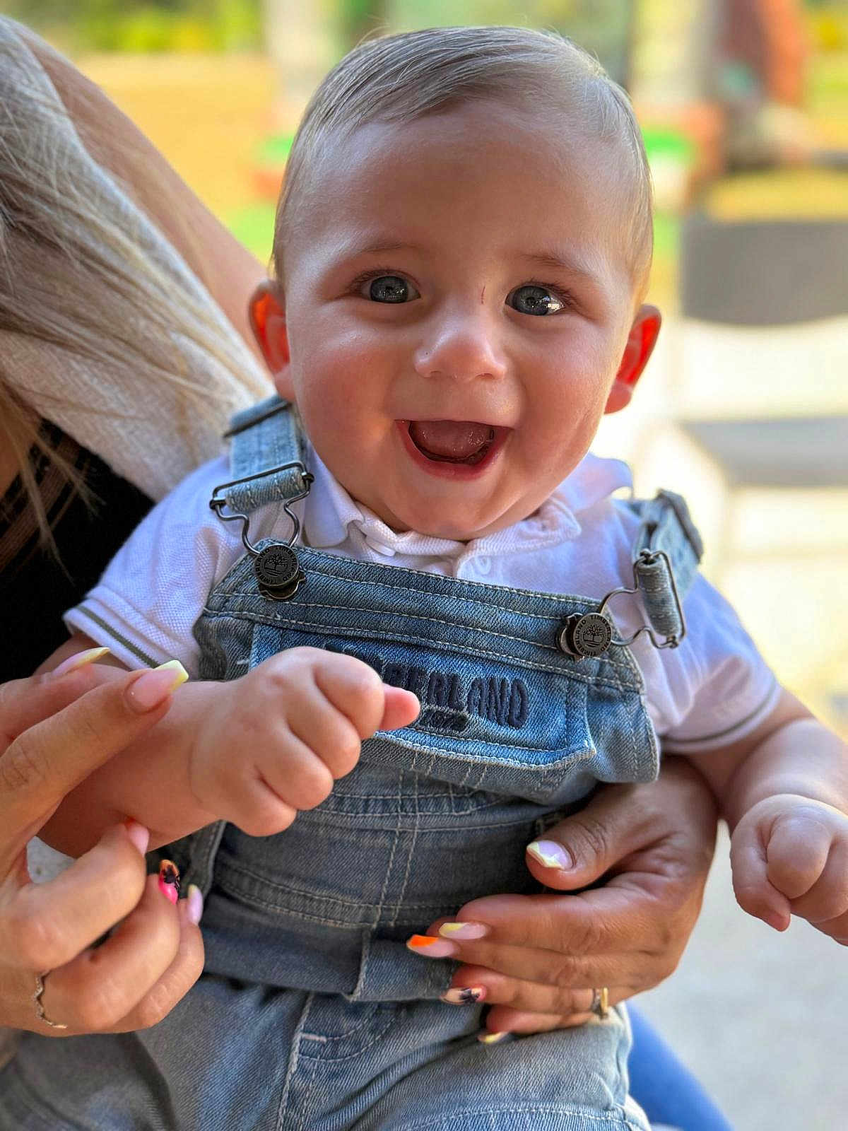 Tao a rejoint le concours — aidez-le/la à gagner de superbes lots ! baby, child, smiling, happy, denim_overalls, white_shirt, hands, adult, nails, colorful_nail_polish, face, portrait, outdoor, blurred_background, cute, infant, holding, joyful, closeup, person