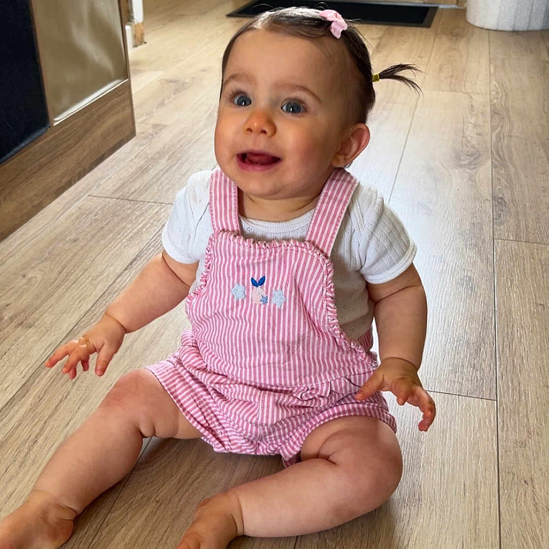 Olivia a rejoint le concours — aidez-le/la à gagner de superbes lots ! baby, child, coat, cute, door, flooring, furniture, hair_bow, happy, home_interior, indoor, jacket, person, pigtails, pink_overalls, playful, sitting, smiling, white_shirt, wooden_floor