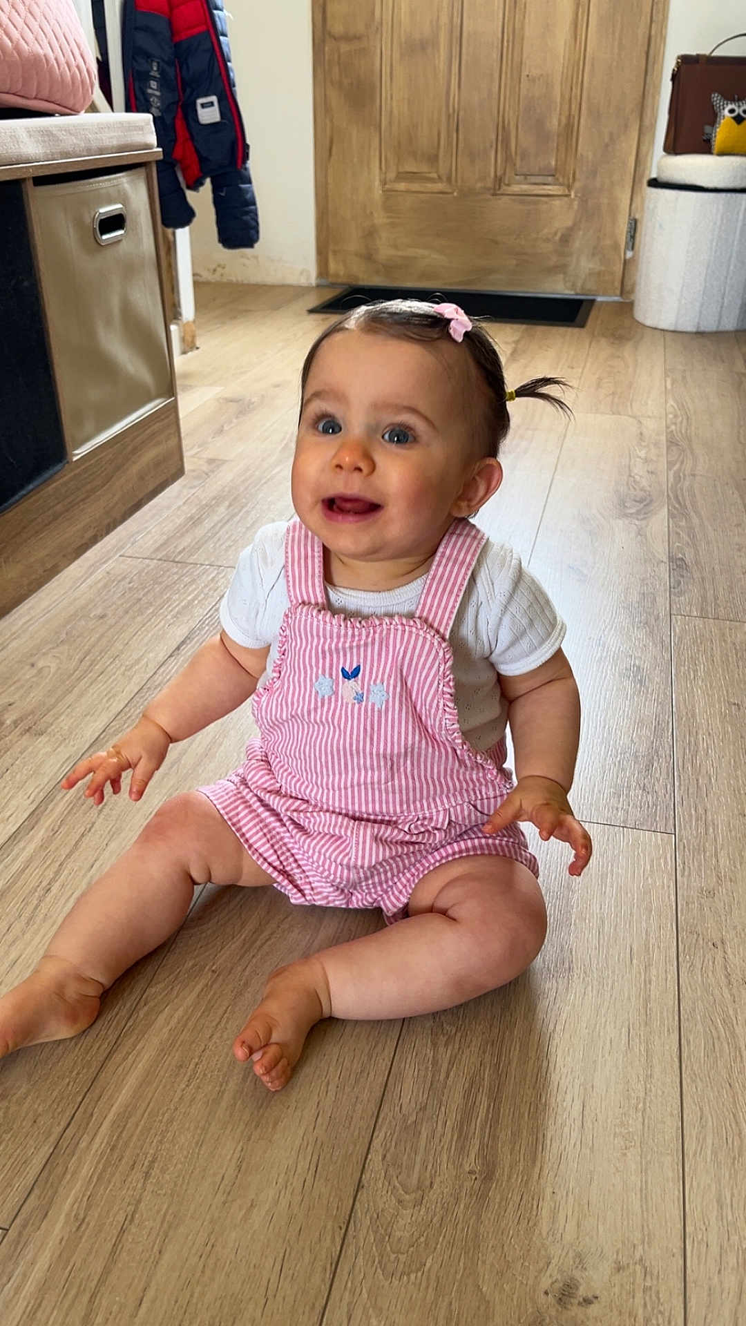 Olivia a rejoint le concours — aidez-le/la à gagner de superbes lots ! baby, child, pink_overalls, white_shirt, hair_bow, pigtails, wooden_floor, door, indoor, smiling, sitting, cute, happy, person, flooring, home_interior, jacket, coat, furniture, playful