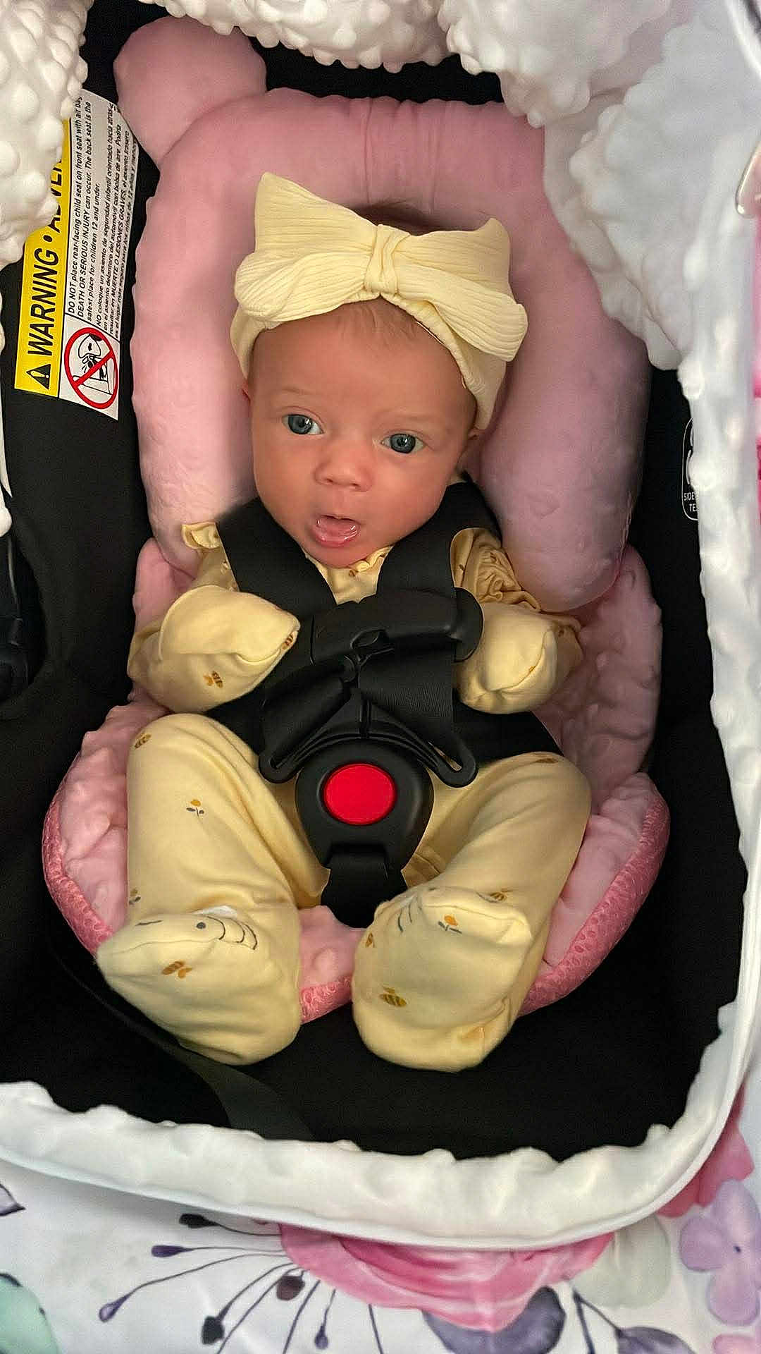 Nala is registered to the contest to win money with this photo: baby, infant, newborn, car_seat, carseat_harness, headband, bow, pink_pillow, onesie, footed_pajamas, buckle, red_button, seat_padding, warning_label, portrait, wide_eyes, open_mouth, floral_blanket, soft_lighting, cute