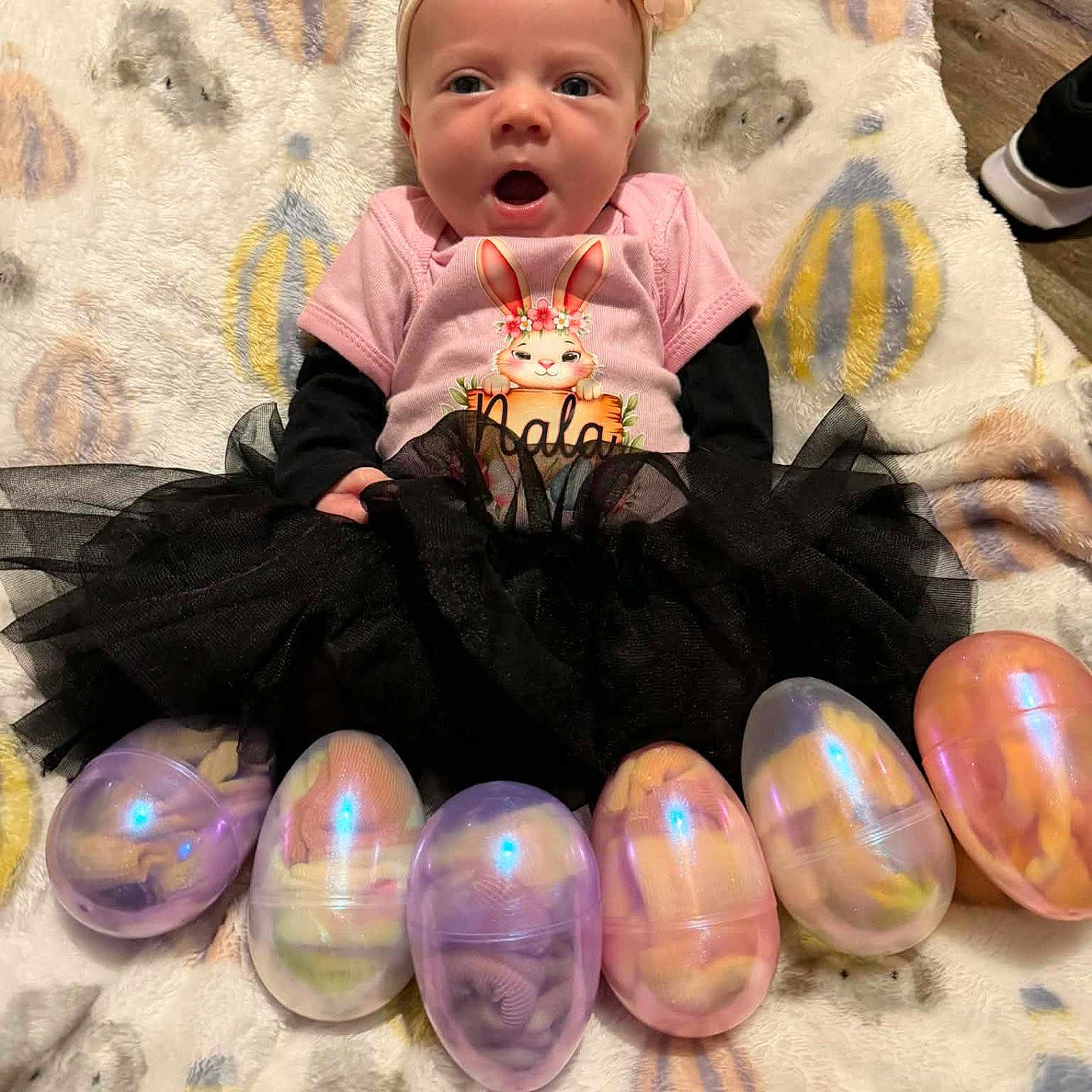 Nala joined the competition — help win amazing prizes! baby, infant, child, girl, headband, flower, pink_shirt, bunny_graphic, black_tutu, plastic_eggs, easter_eggs, blanket, patterned_blanket, floor, toy, cute, lying_down, indoors, colorful, smiling
