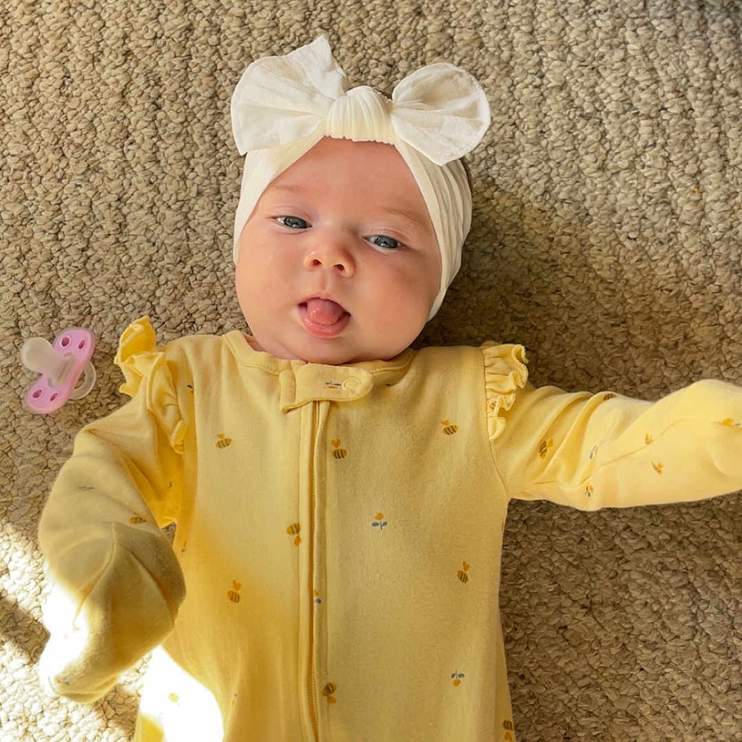 Nala joined the competition — help win amazing prizes! baby, infant, child, headband, bow, yellow_onesie, carpet, pacifier, tongue_out, cute, playful, indoor, soft_light, texture, lying_down, cozy, adorable, smiling, person, young_child
