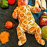 baby, taco_onesie, infant, food, vegetables, cheese, onion, bell_pepper, tomato, lettuce, tortilla, taco_sauce, towel, laying_down, headband, cute, playful, indoor, portrait, colorful