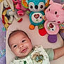 baby, infant, smiling, plush_toys, colorful, cute, floral_outfit, decorative_pin, play_area, comfort, lying_down, soft_toys, pastel_colors, happy, child, face, portrait, indoors, young_child, toy