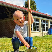 Augustin a rejoint le concours — aidez-le/la à gagner de superbes lots ! toddler, child, outdoor, grass, garden_tool, blue_sky, clouds, building, window, sunny, casual_clothing, jeans, short_sleeve_shirt, greenery, daytime, playful, crouching, nature, young_child, summer