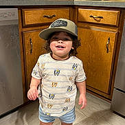 Aiden joined the competition — help win amazing prizes! toddler, child, smiling, hat, baseball_cap, kitchen, wooden_cabinets, floor, shorts, tshirt, barefoot, standing, home, indoors, pot, cup, countertop, appliance, person, casual_clothing