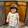 toddler, child, smiling, hat, baseball_cap, kitchen, wooden_cabinets, floor, shorts, tshirt, barefoot, standing, home, indoors, pot, cup, countertop, appliance, person, casual_clothing
