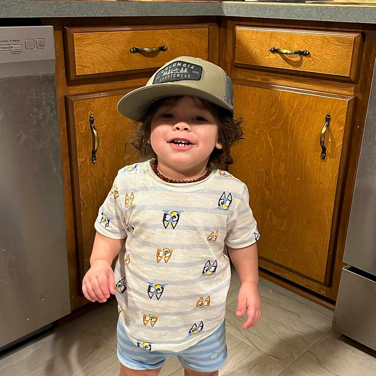 Aiden joined the competition — help win amazing prizes! toddler, child, smiling, hat, baseball_cap, kitchen, wooden_cabinets, floor, shorts, tshirt, barefoot, standing, home, indoors, pot, cup, countertop, appliance, person, casual_clothing