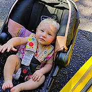 Emery is registered to the contest to win money with this photo: baby, child, car_seat, harness, blonde_hair, outdoor, pavement, yellow_curb, clothing, seat_belt, infant, person, cute, sitting, foot, hand, sunlight, colorful, striped, safety